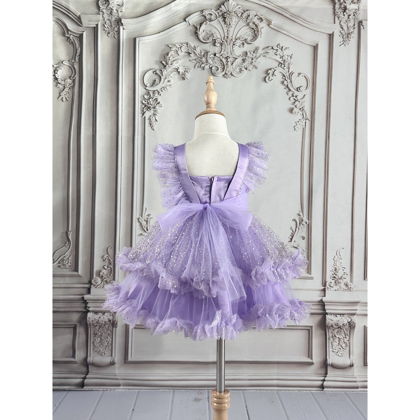 A light purple girl's dress with delicate butterfly patterns at the top, silver linear decor throughout the dress, and purple tulle, displayed on a wooden stand with an ornate white background, back side