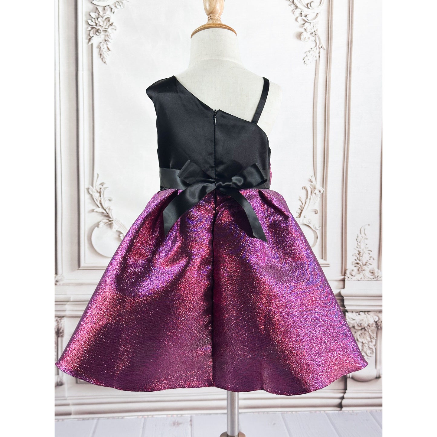 A metallic purple bow dress with a metallic purple bow, black at the top, thick sleeve on left, thin shoulder strap on the right, displayed on a wooden stand in with a elegant background, back view