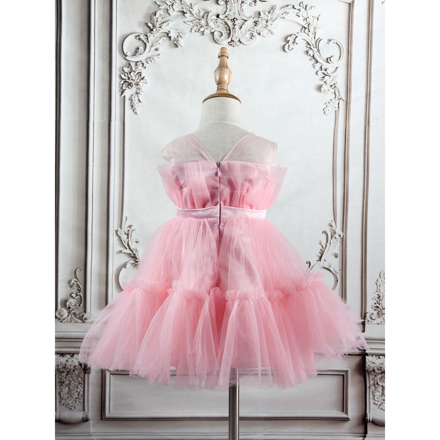 A pink tulle princess dress displayed on a mannequin stand, featuring a pleated top, bow belt at the center, and sleeveless, image is of the back of the dress
