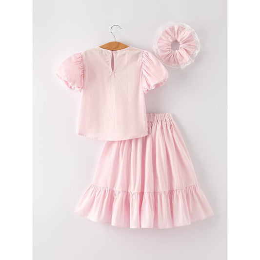 Pink outfit with top that has bubble sleeves and skirt that has elastic waist and ruffles at the bottom of skirt on a hanger, accompanied by a matching hair scrunchie with white lace around on a light gray background. Easter dress, spring dress Inso, lightweight material, easy wear