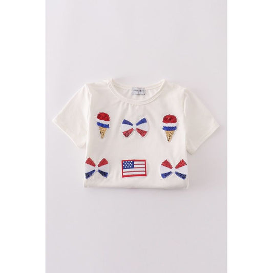 White t-shirt with embroidered ice cream cones, bows, and flags on a light gray background. ice cream, bows and flag have sequins in red white and blue for 4th of july
