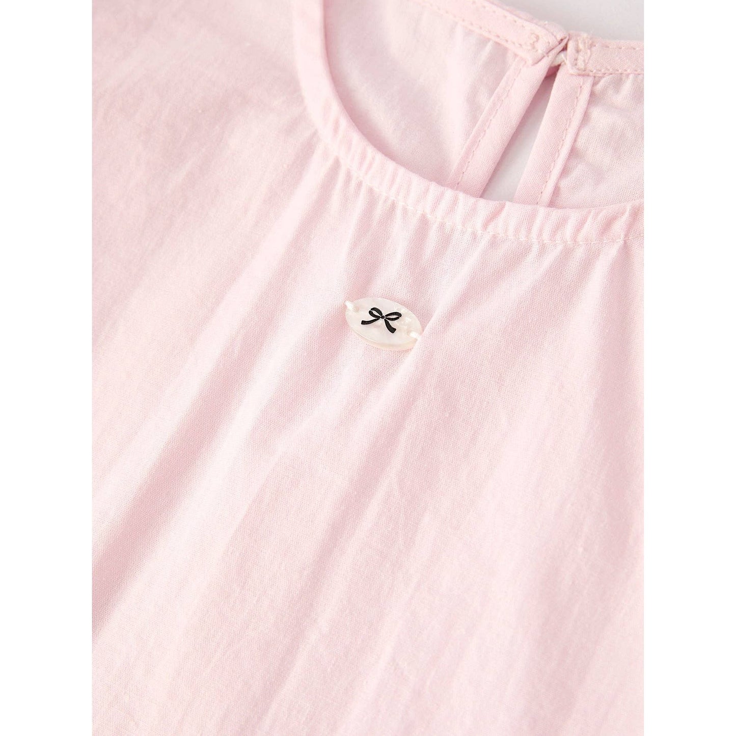 Pink outfit with top that has bubble sleeves and skirt that has elastic waist and ruffles at the bottom of skirt on a hanger, accompanied by a matching hair scrunchie  with white lace around on a light gray background. close up of small black bow design button sewed to front of top