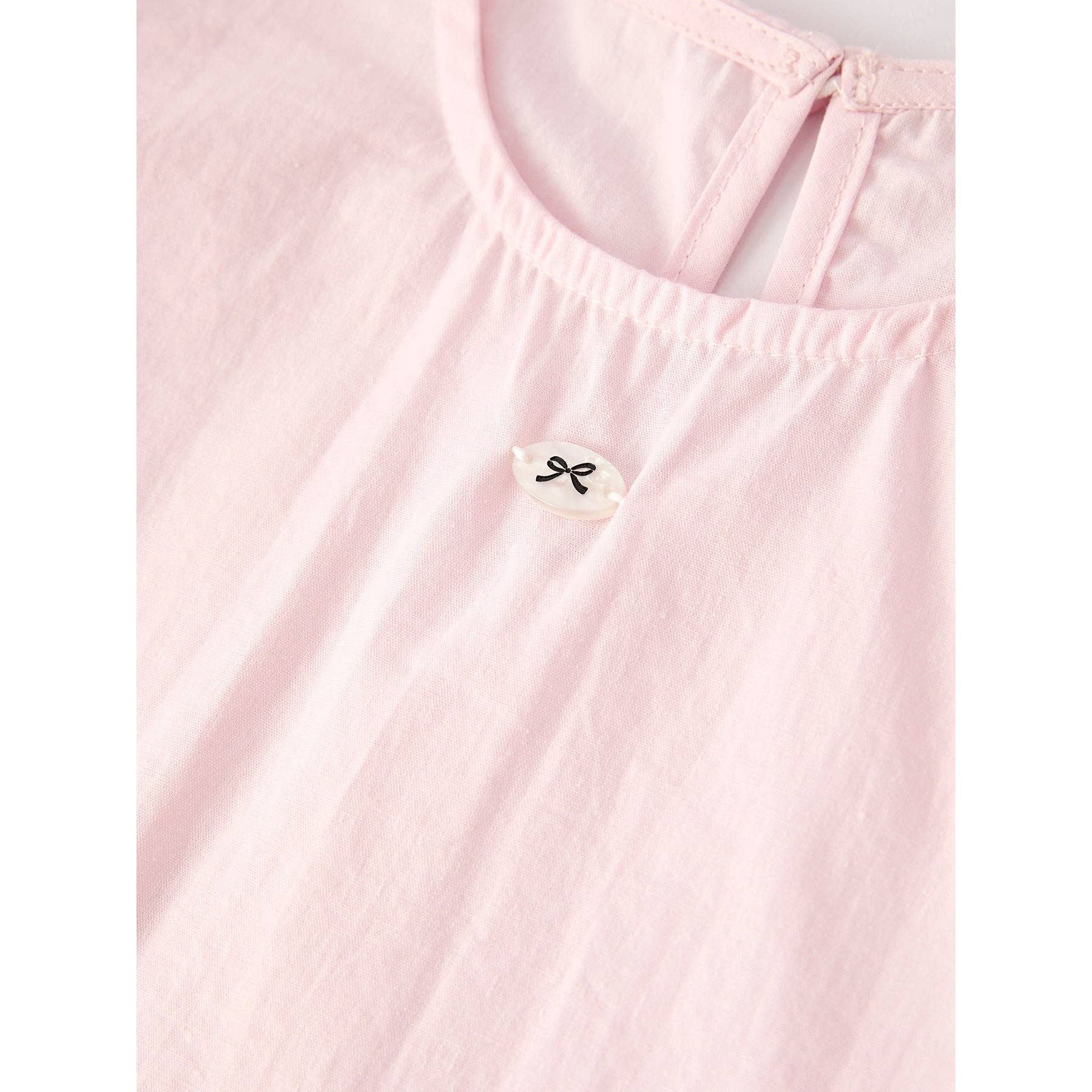 Pink outfit with top that has bubble sleeves and skirt that has elastic waist and ruffles at the bottom of skirt on a hanger, accompanied by a matching hair scrunchie  with white lace around on a light gray background. close up of small black bow design button sewed to front of top