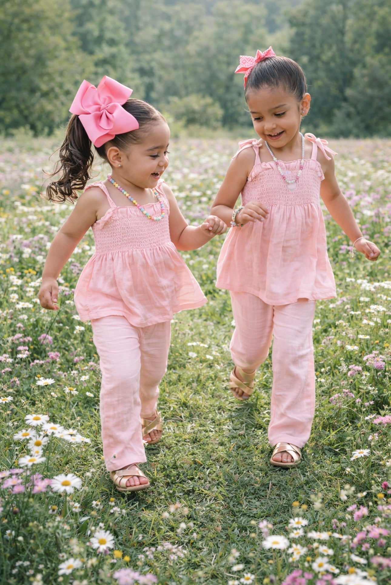 Two young girls in matching pink outfits walking through a field of flowers.