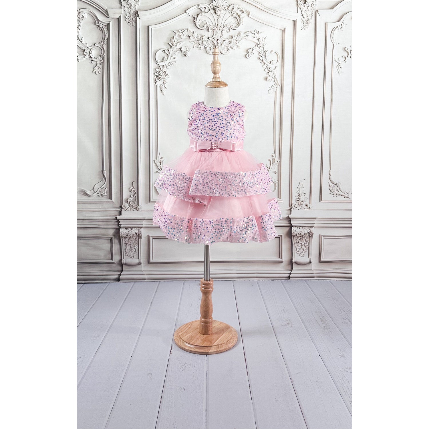 A light pink tulle dress with sequins and a bow belt, displayed on a wooden stand with an elegant neutral colored background.