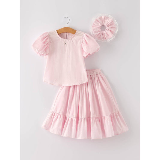 Pink outfit with top that has bubble sleeves and skirt that has elastic waist and ruffles at the bottom of skirt on a hanger, accompanied by a matching hair scrunchie  with white lace around on a light gray background.
